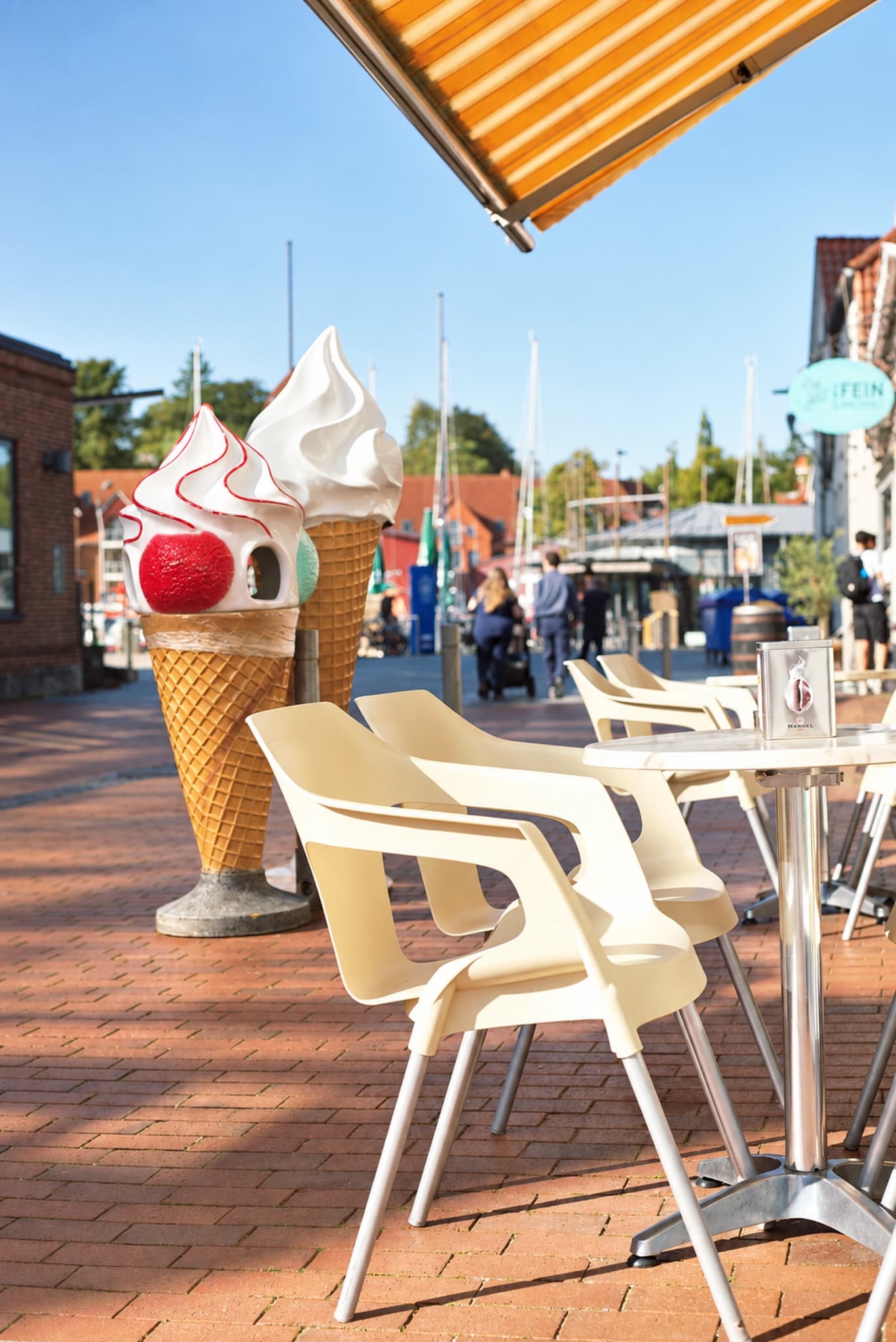 Zwei große Eistüten-Figuren und weiße Stühle vor dem Eiscafé am Hafen, im Hintergrund Bootsmasten