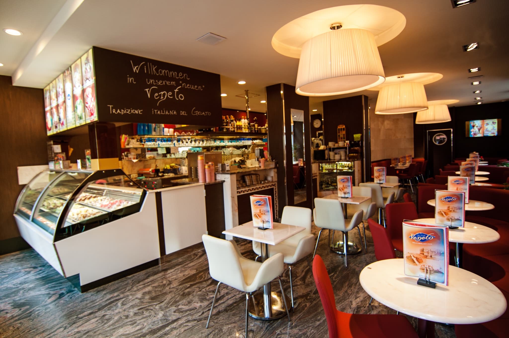 Innenraum von Eiscafé Veneto mit gewölbter Eis-Vitrine, der Kreidetafel „Willkommen in unserem ‚neuen‘ Veneto – Tradizione italiana del Gelato“ und Marmortischen unter großen Stoff-Hängelampen