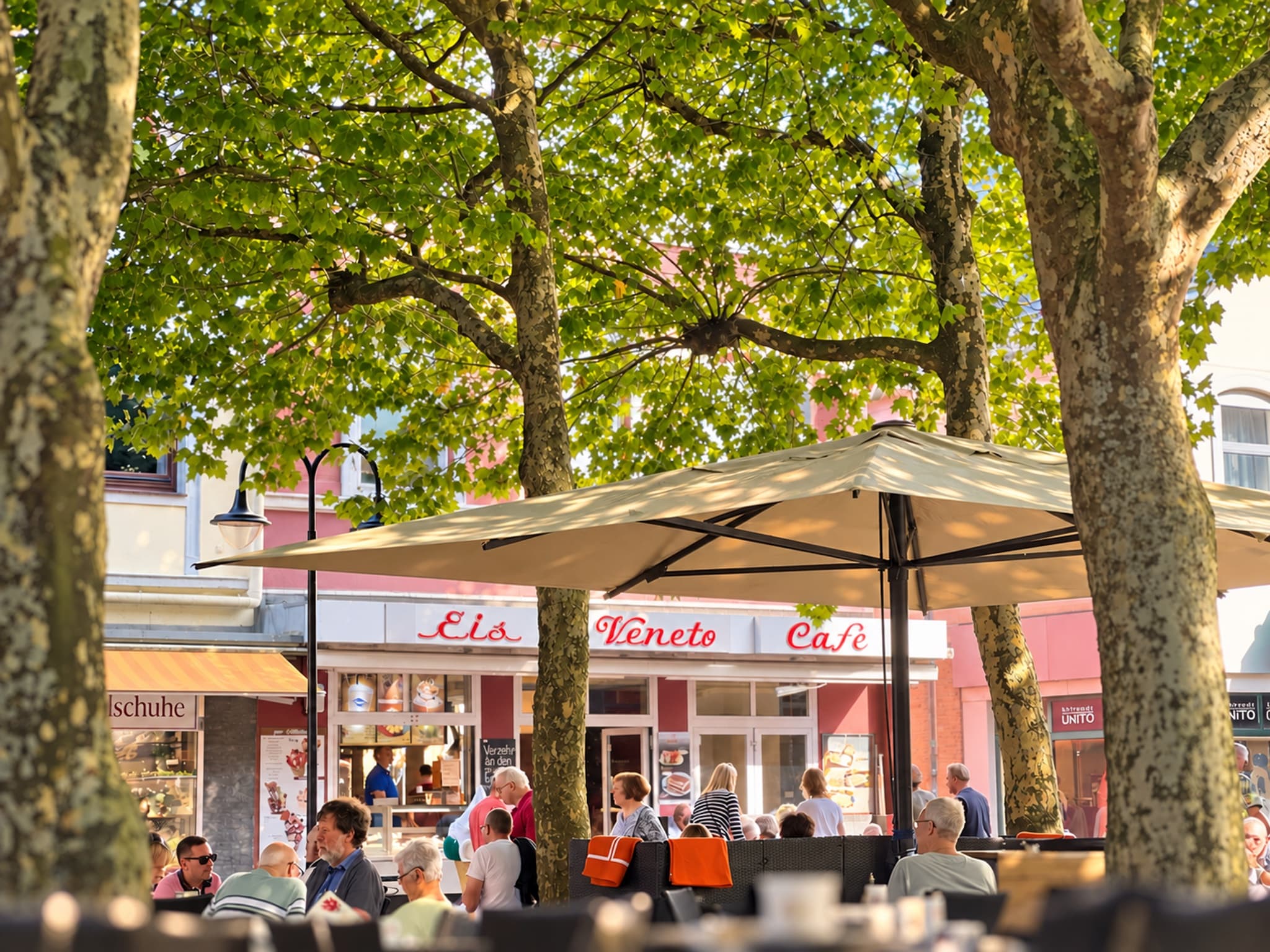 Außenterrasse von Eiscafé Veneto in der Einkaufsmeile unter großen Platanen: Gäste an den Tischen, dahinter die rote Fassade mit dem Schriftzug „Eis Veneto Café“