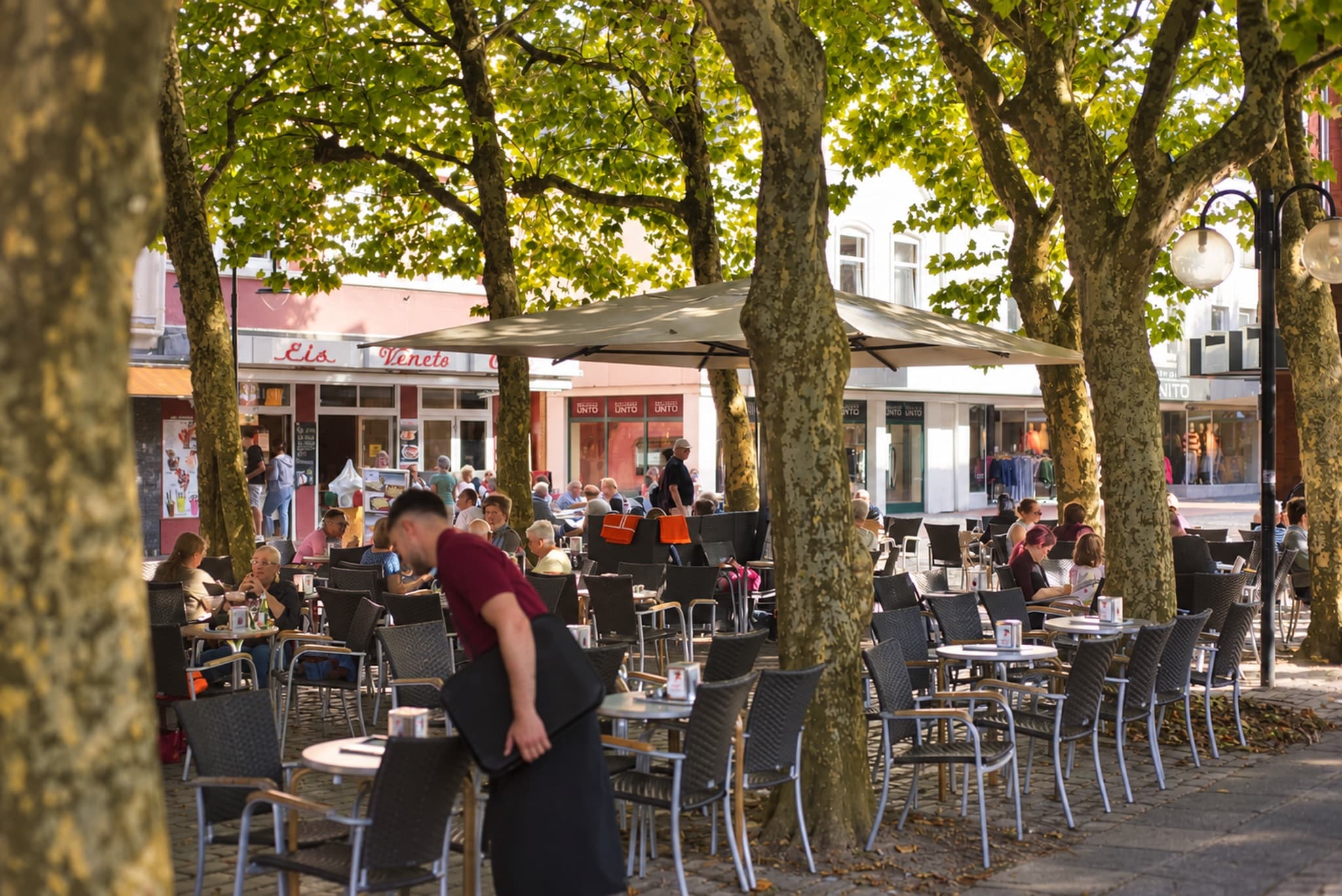 Terrasse in der Einkaufsmeile unter Platanen: ein Mann im roten Poloshirt beugt sich über einen Tisch, im Hintergrund die rote Veneto-Fassade