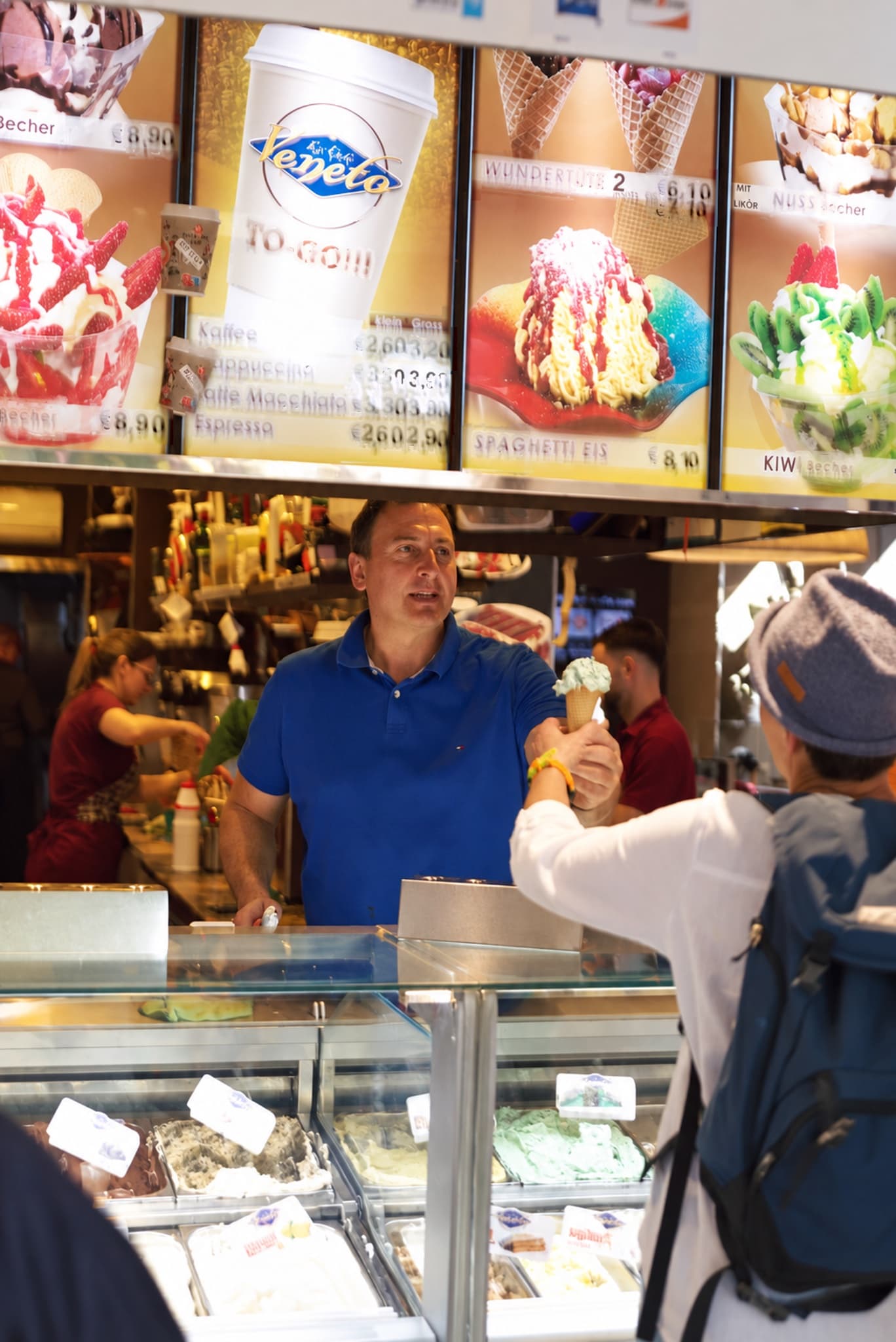 In der Einkaufsmeile reicht ein Mitarbeiter im blauen Poloshirt einem Gast eine Eiswaffel über die Vitrine, darüber beleuchtete Bildtafeln mit Eisbechern